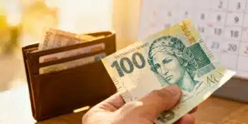 Mão segurando uma nota de cem reais com carteira ao fundo, simbolizando o Abono Salarial PIS/PASEP 2026.